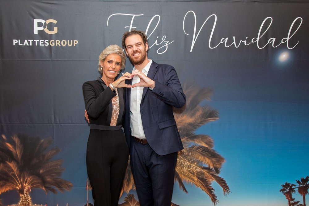Die Geschäftsführerin der PlattesGroup, Yvonne Plattes, und ihr Sohn Christian Plattes bilden mit ihren Händen ein Herz. Sie sind festlich für die Weihnachtsfeier gekleidet und posieren vor einer Fotowand mit dem Schriftzug "Feliz Navidad".